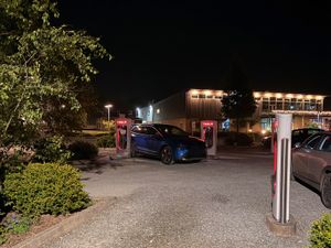 Wide nighttime view of a blue Skoda Elroq 80 charging at a Tesla Supercharger station, surrounded by several empty stalls with their red cables hanging ready, in a quiet gravel car park with a lit building in the background