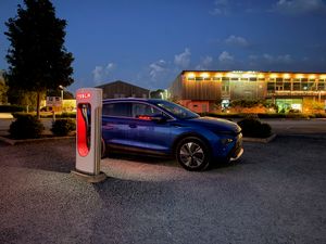 A blue Skoda Elroq 80 plugged into a Tesla Supercharger at dusk, its red charging port glowing against the car's metallic paint, with the warmly-lit Darts Farm retail village in Exeter visible in the background