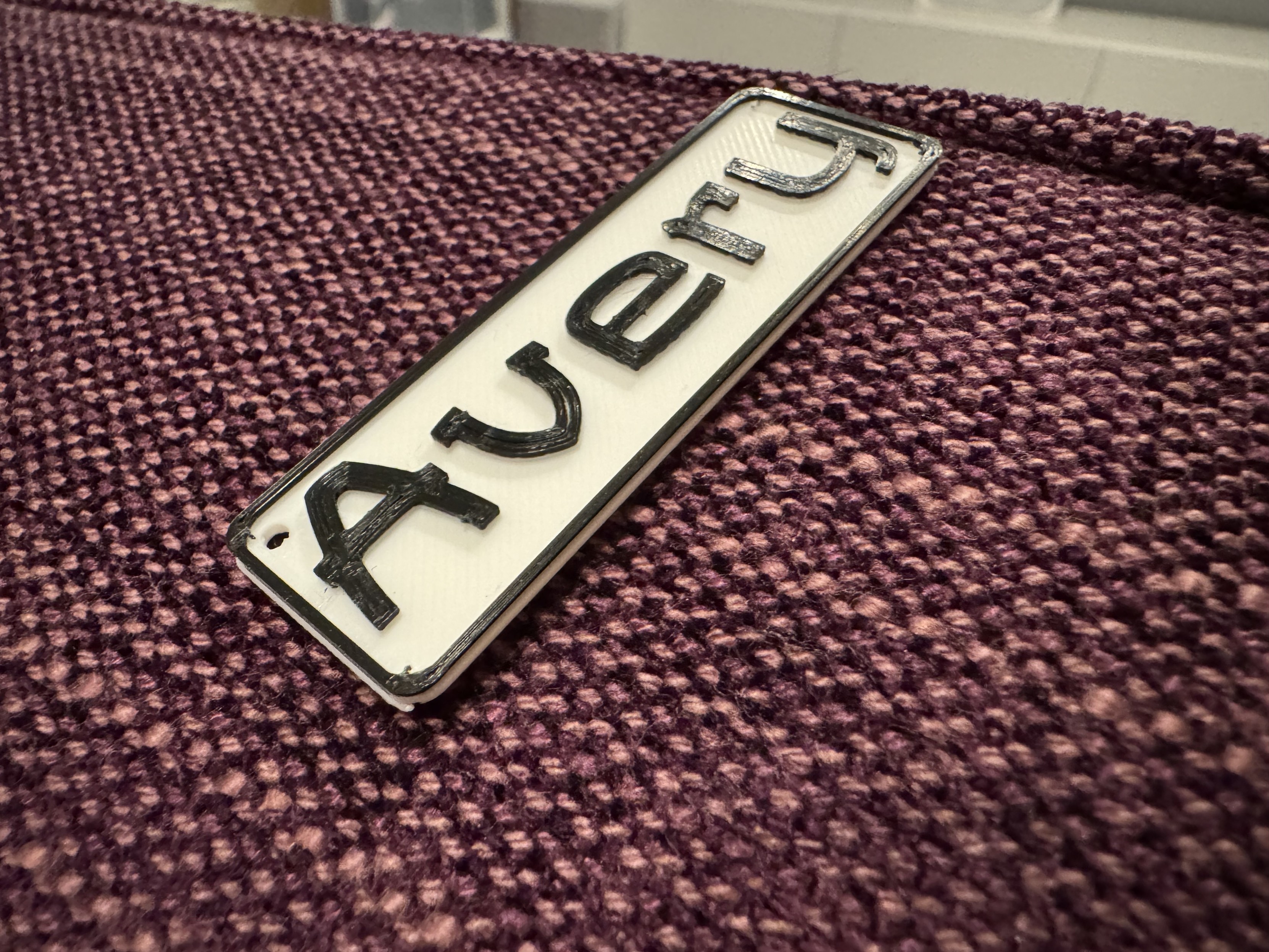 A keyring with the name "Avery" in raised black letters on a white background, lying on a textured purple fabric surface.