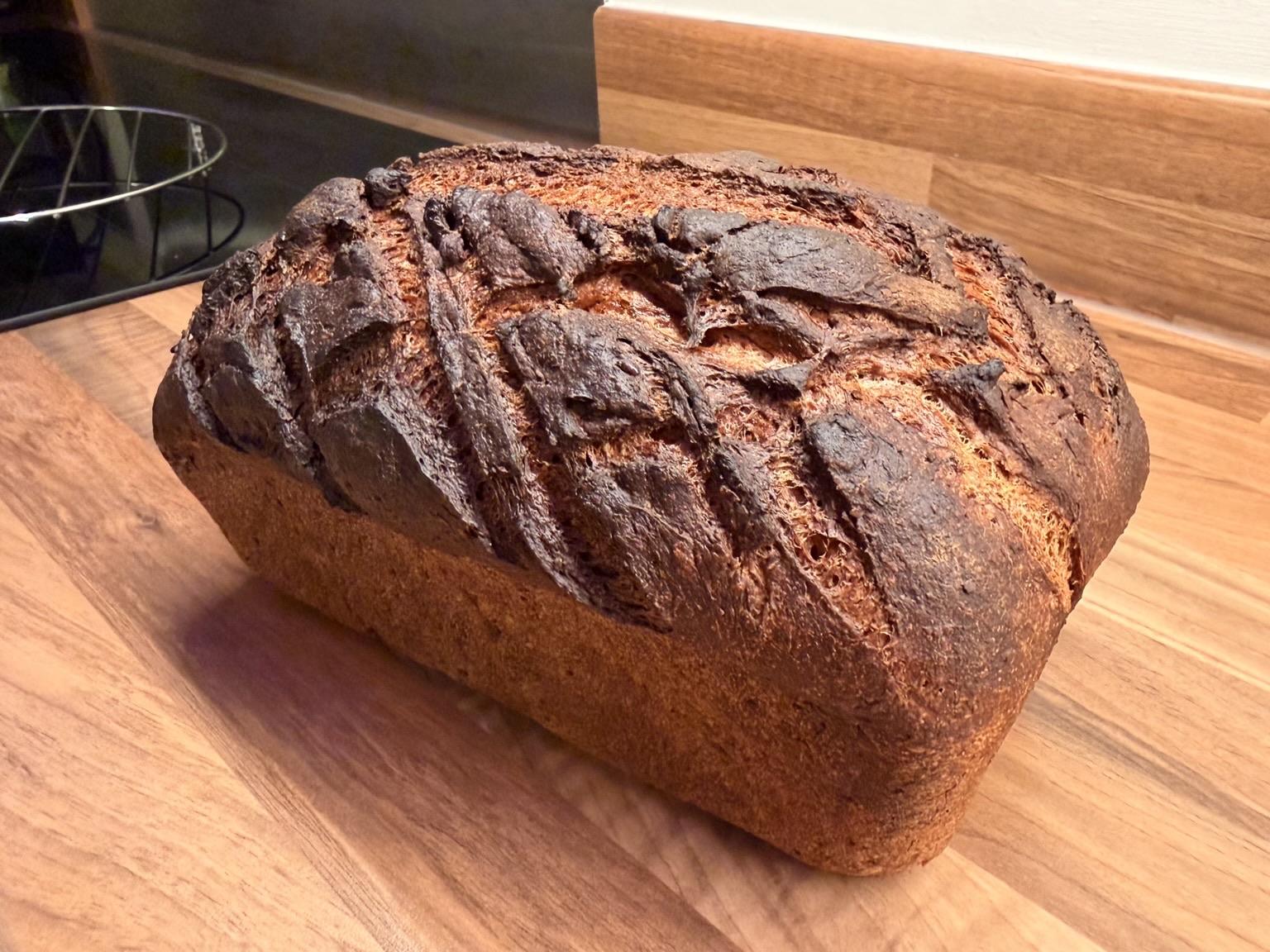 A freshly baked loaf of dark brown bread with a textured crust, sitting on a wooden surface.