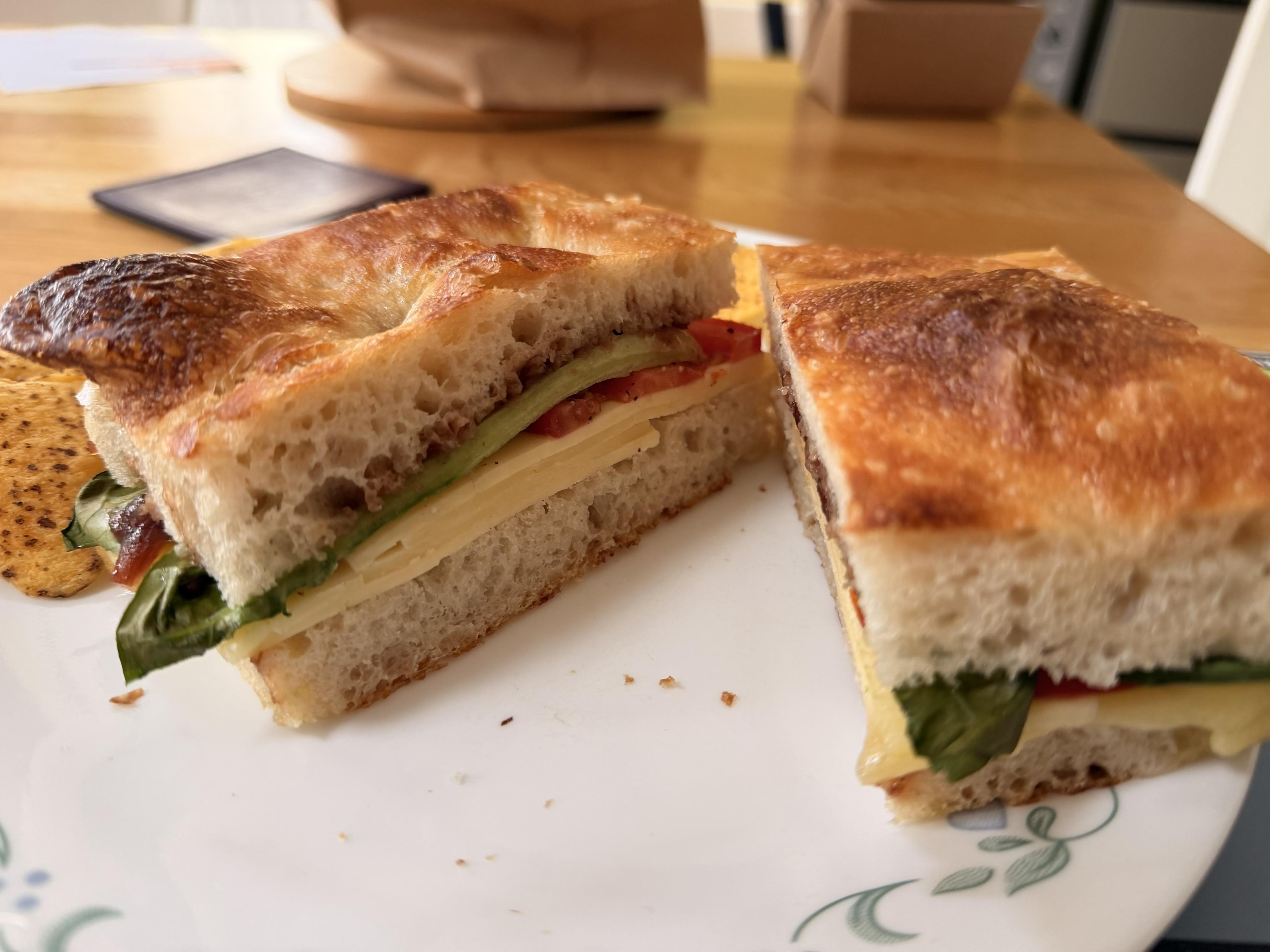 A close-up of a sliced sandwich on a plate, showcasing layers of cheese, leafy greens, and sliced vegetables. The bread is crusty and golden-brown. A few crisps are visible on the side.