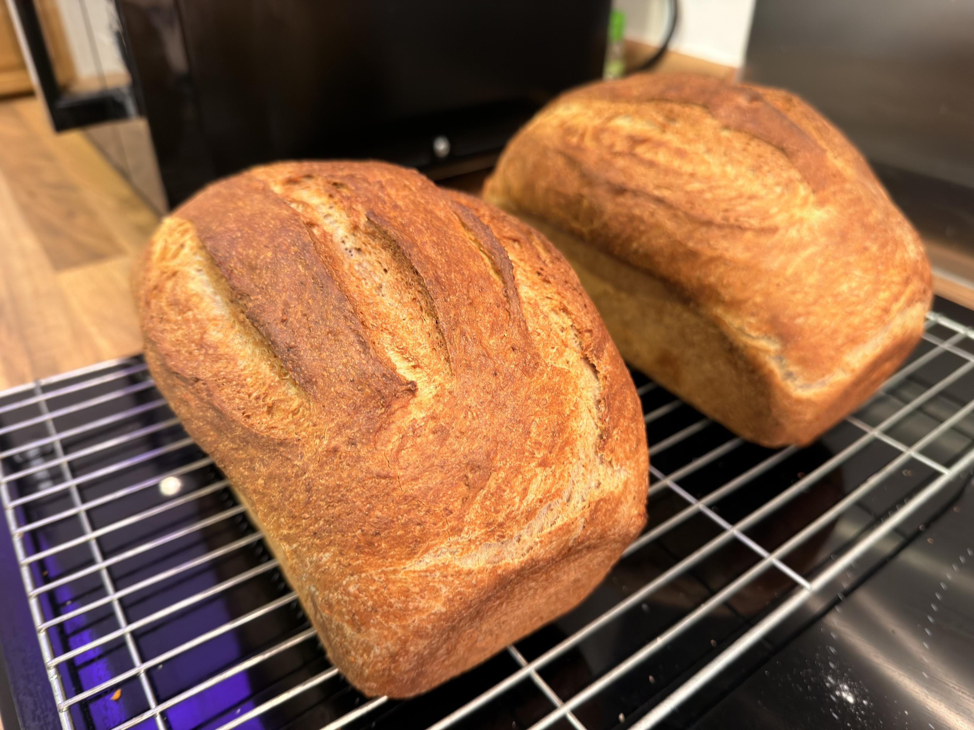 Two freshly baked loaves of brown bread cooling on a wire rack. The bread has a golden-brown crust with visible scoring on top. Background features a dark kitchen setting.