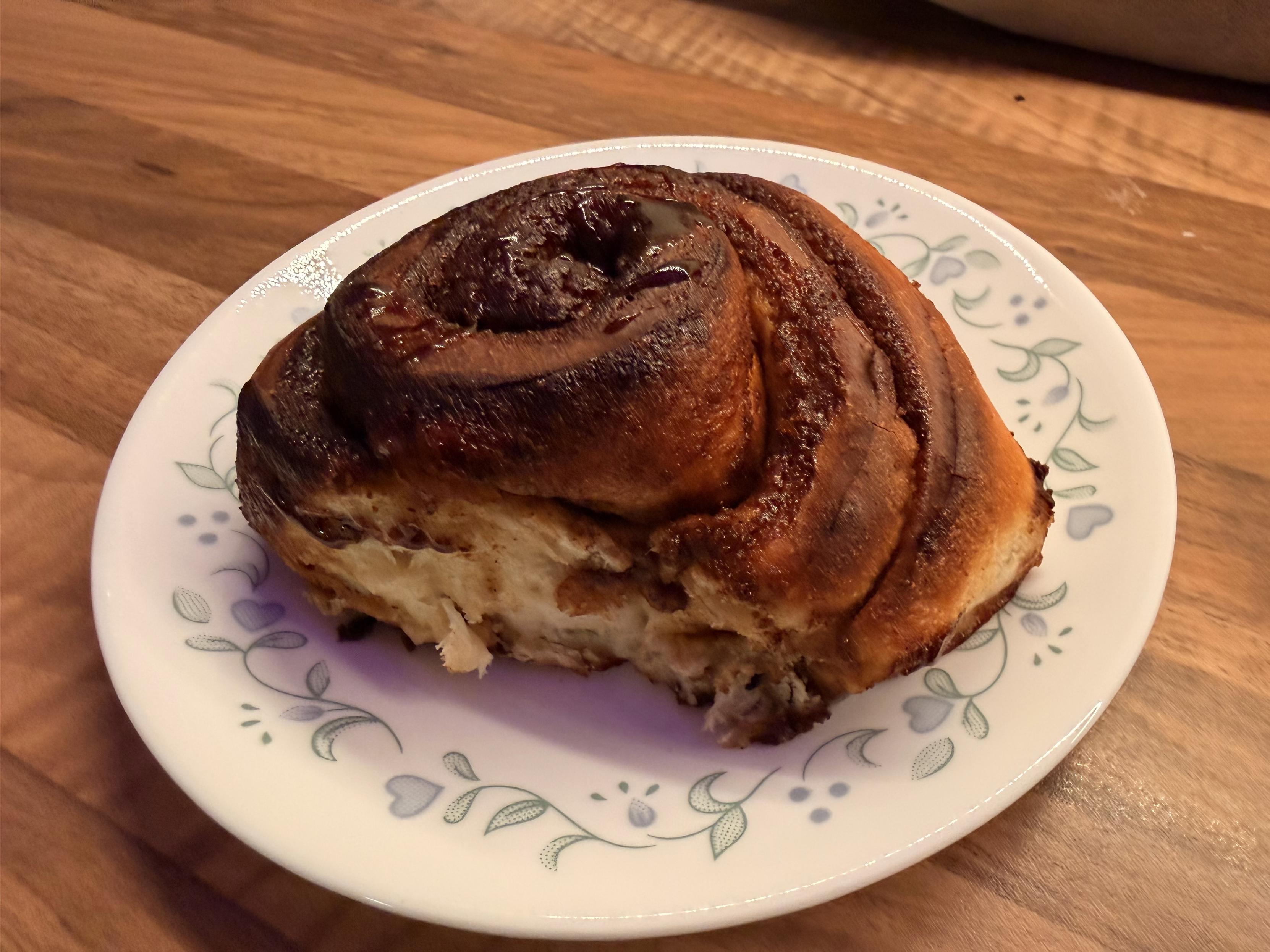 A freshly baked cinnamon roll on a decorative plate, resting on a wooden surface. The roll has a golden-brown exterior with visible swirls of cinnamon.