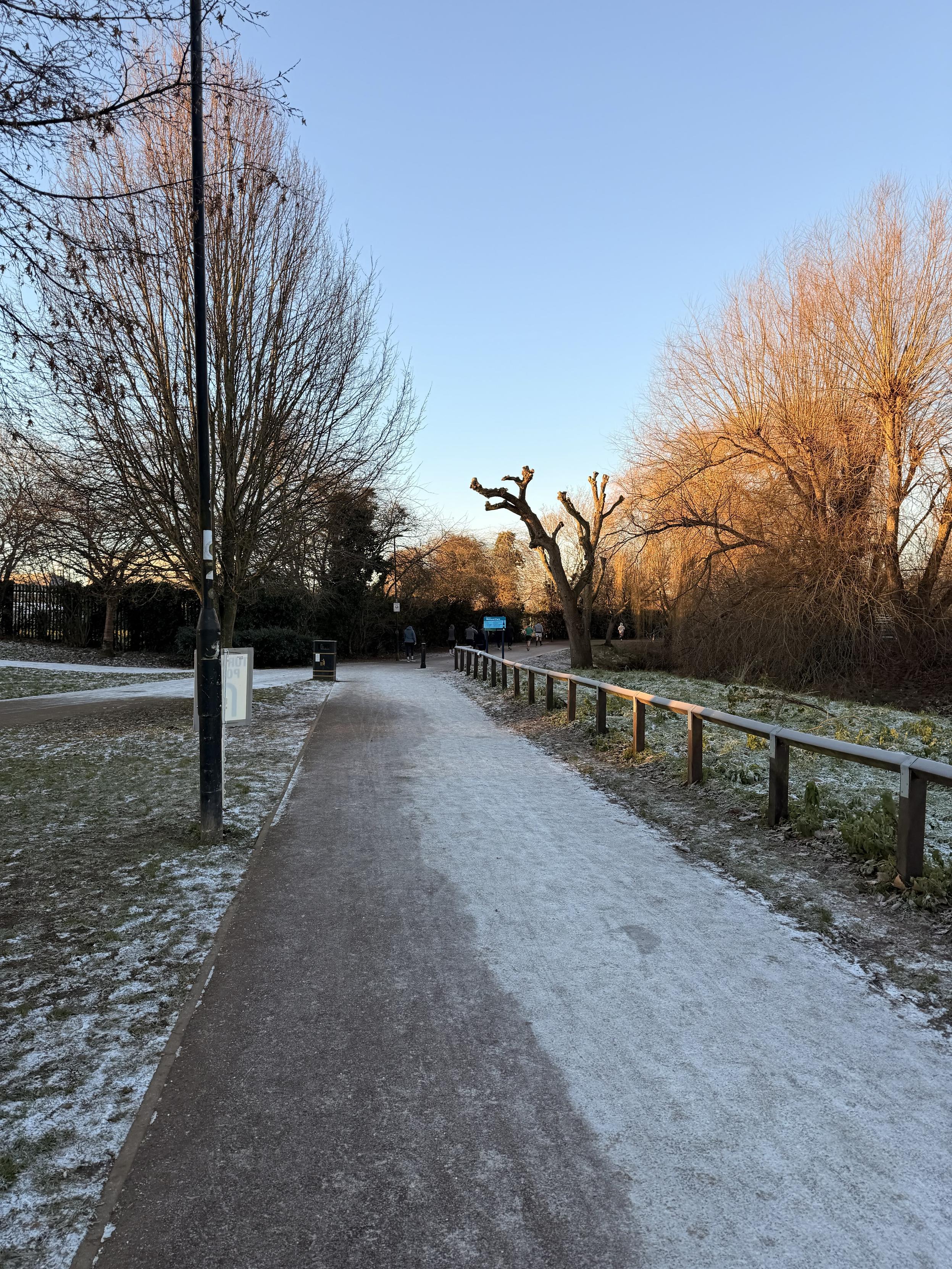 A frost-covered pathway in a park, lined by trees with bare branches. A light post and a wooden railing are visible, with a clear blue sky above. The scene includes hints of green foliage and a few people in the distance.
