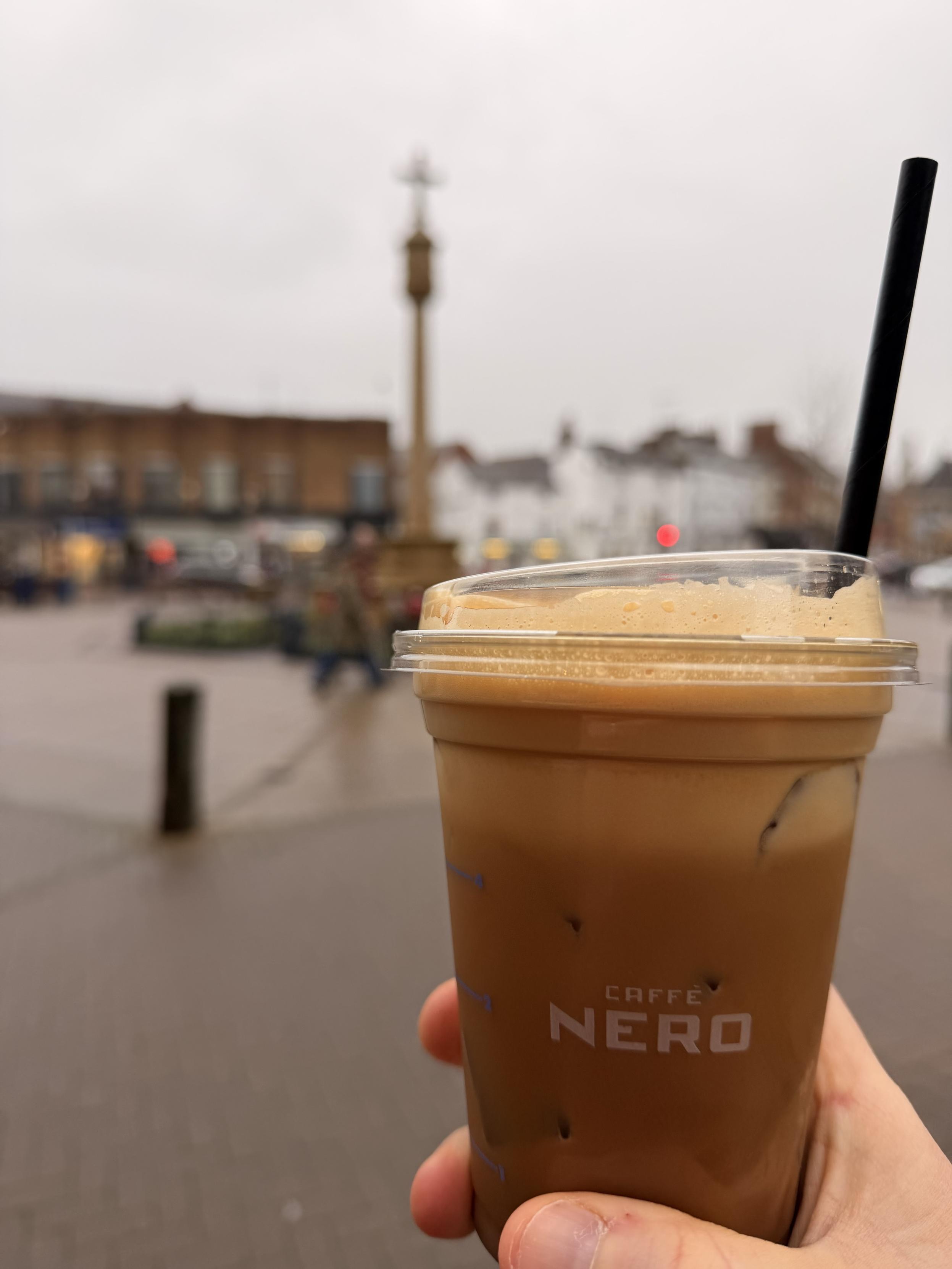 A hand holds a transparent cup of iced coffee with a black straw, set against a blurred background of a town square featuring a monument and distant buildings, under overcast skies.