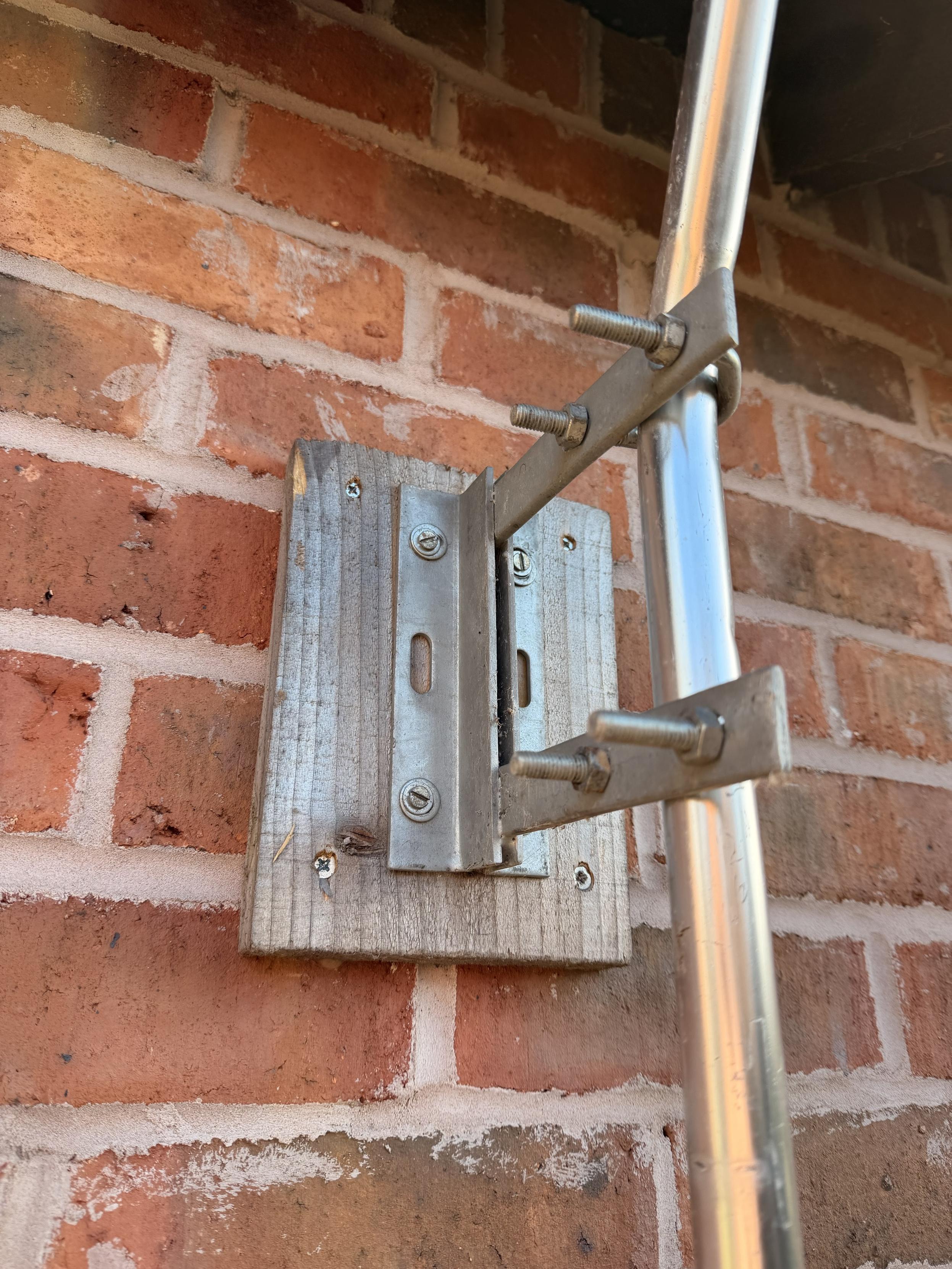 A metal bracket is mounted on a wooden board attached to a red brick wall, supporting a vertical metal pole with screws visible on both the bracket and the wood.