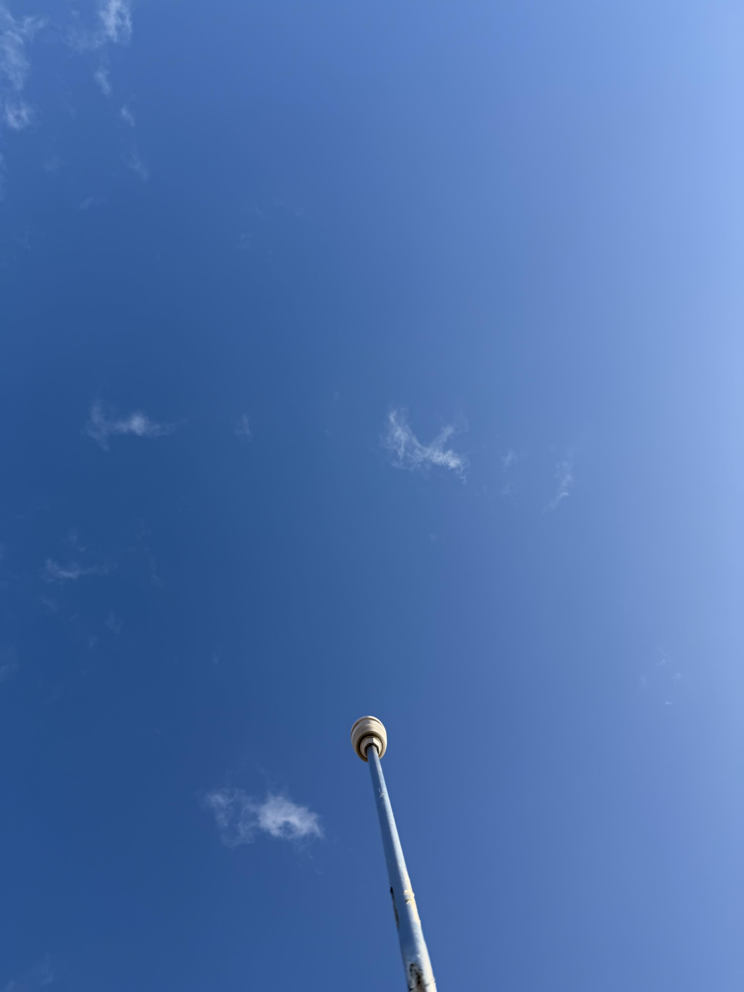 A tall poll with a small white weather station on top reaching towards a clear blue sky, with a few wispy clouds present.