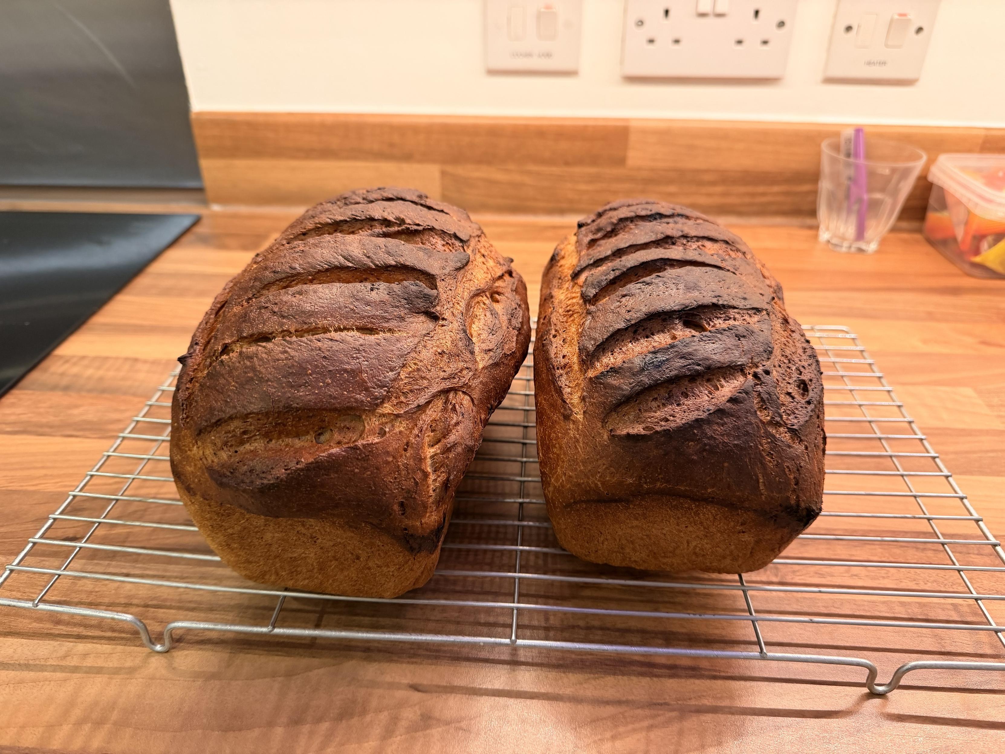 Two loaves of dark, crusty bread cooling on a wire rack against a wooden countertop.
The left loaf is slightly bigger than the right.
The bread has distinct slashes on top and a rich golden-brown color.