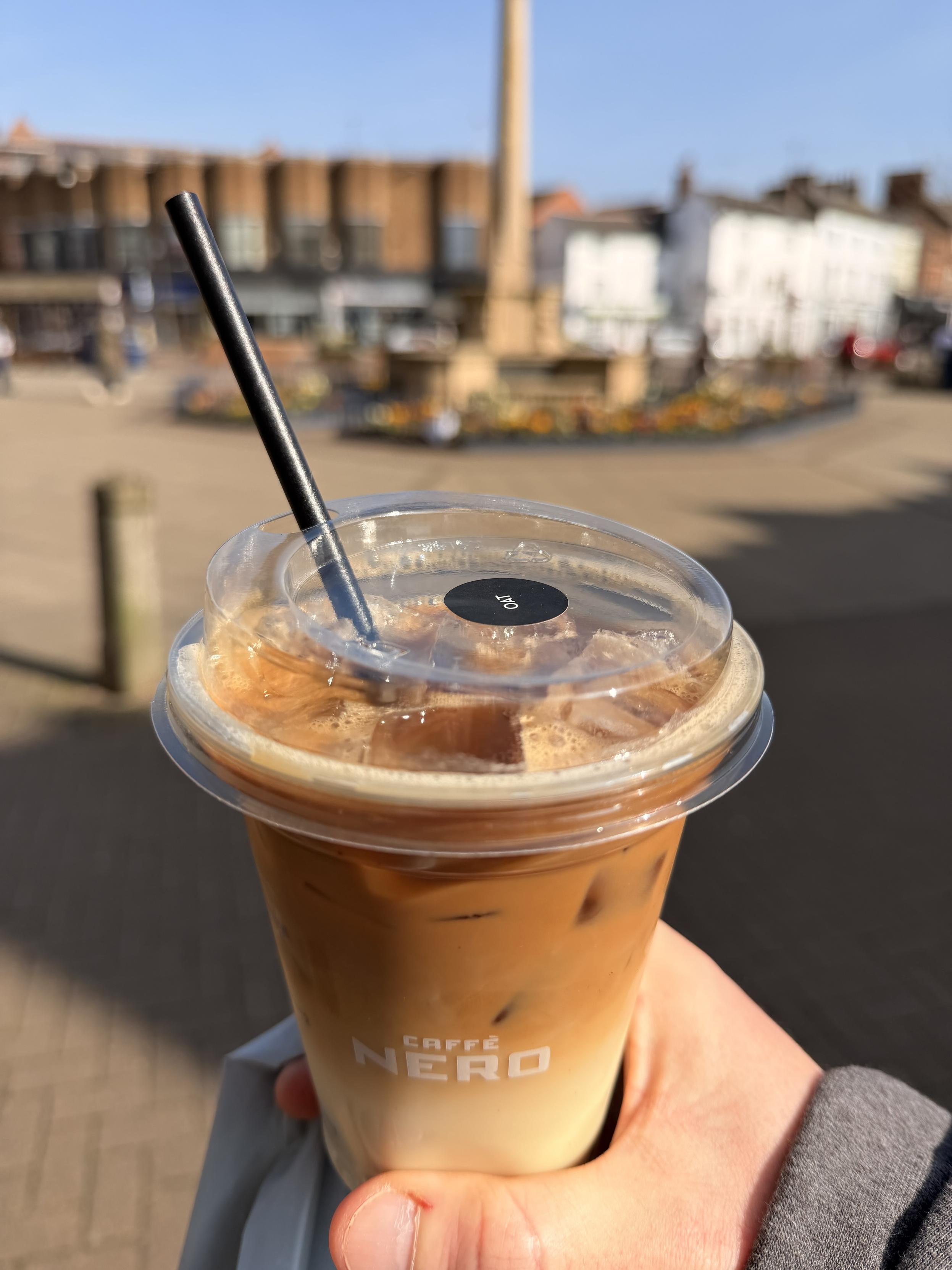 An iced coffee drink with a black straw is held in front of a blurred urban square, featuring a monument and colorful flowerbeds in the background. The cup has a label for "Caffè Nero."