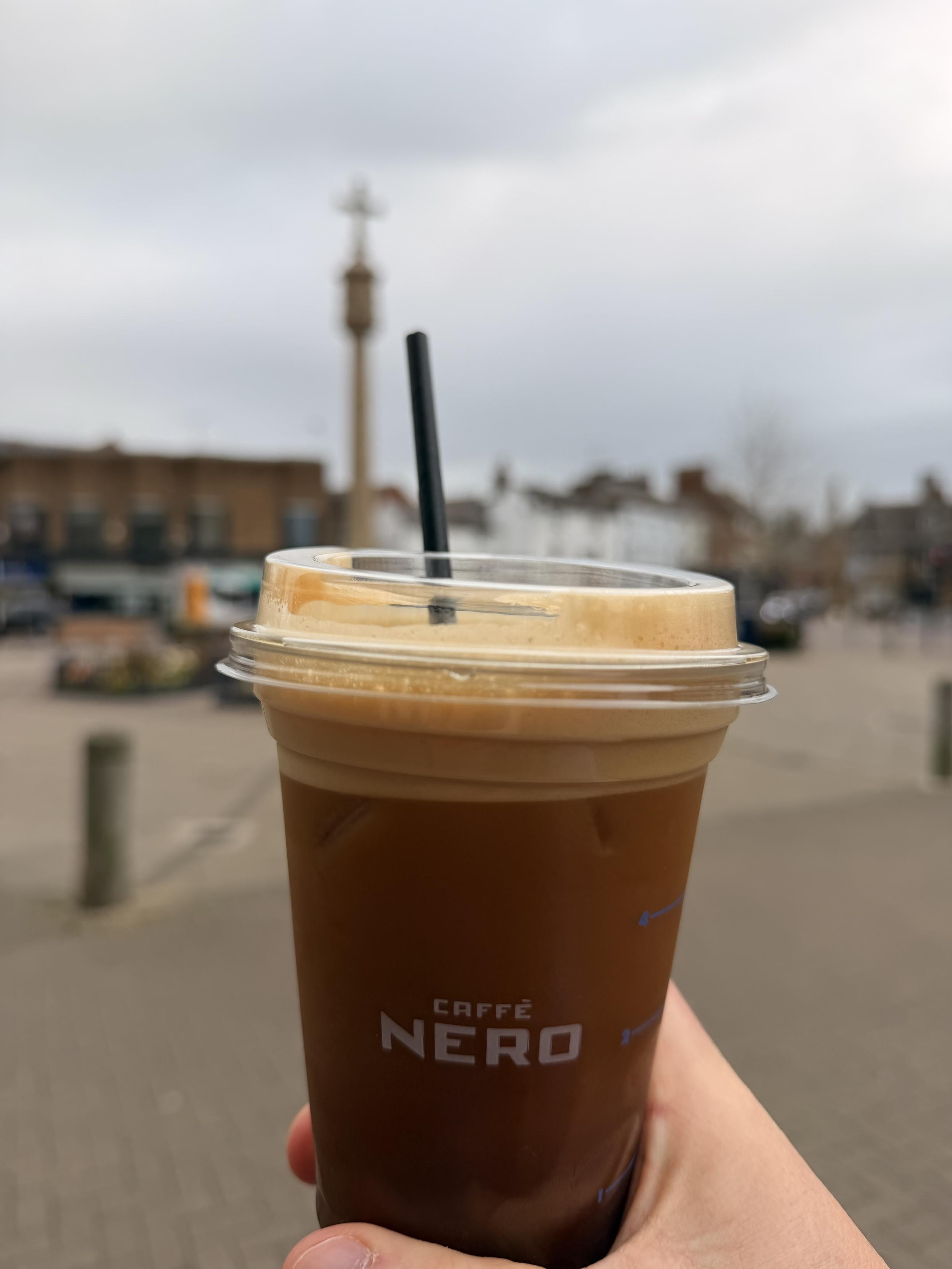 A close-up of an iced coffee drink from Caffè Nero, held in hand, with a black straw. In the background, a monument and buildings can be seen under a cloudy sky.