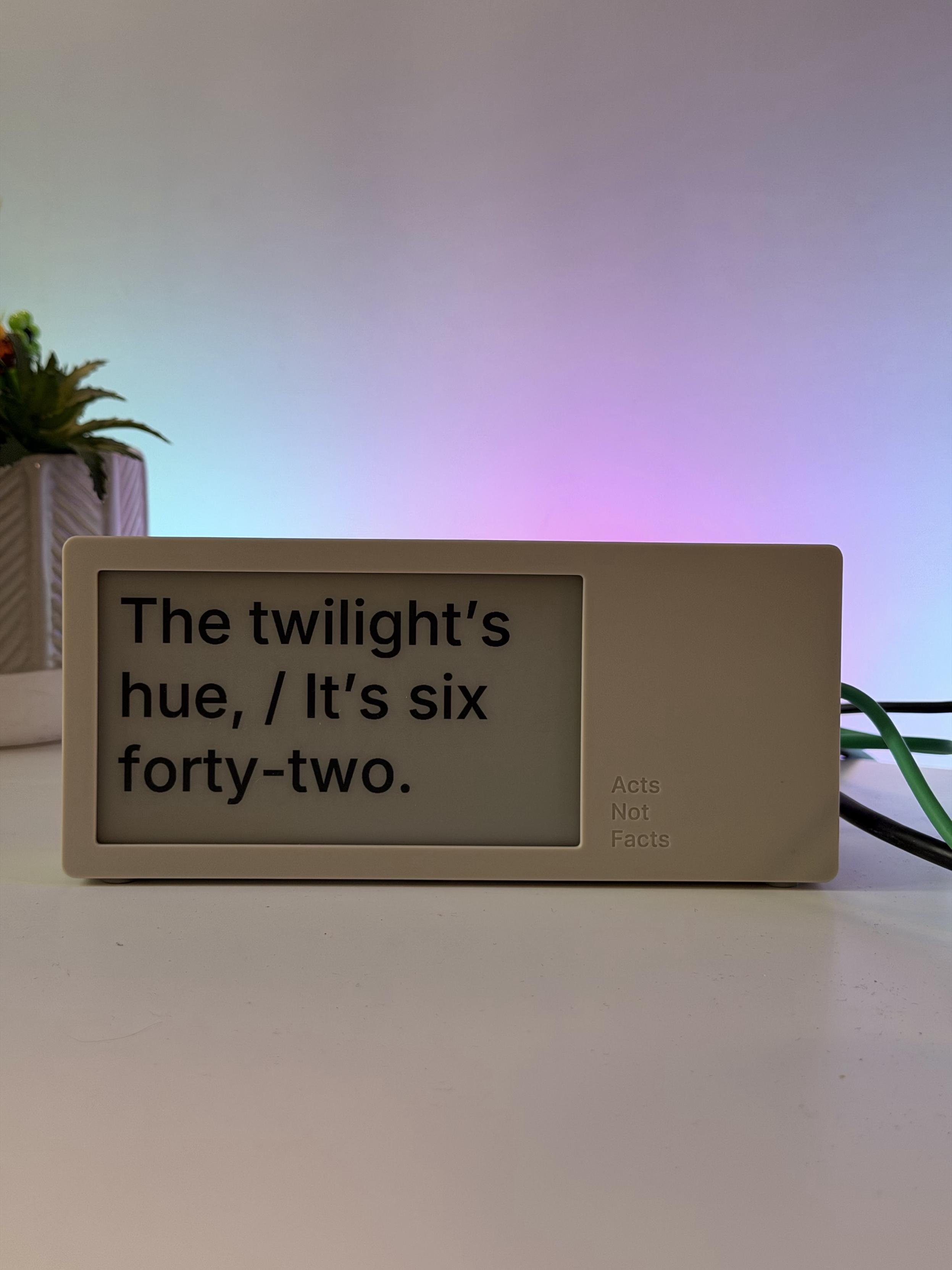 A digital display showing the text: "The twilight's hue, / It's six forty-two." with the phrase "Acts Not Facts" printed on the side. The background features soft, colorful lighting and a potted plant.