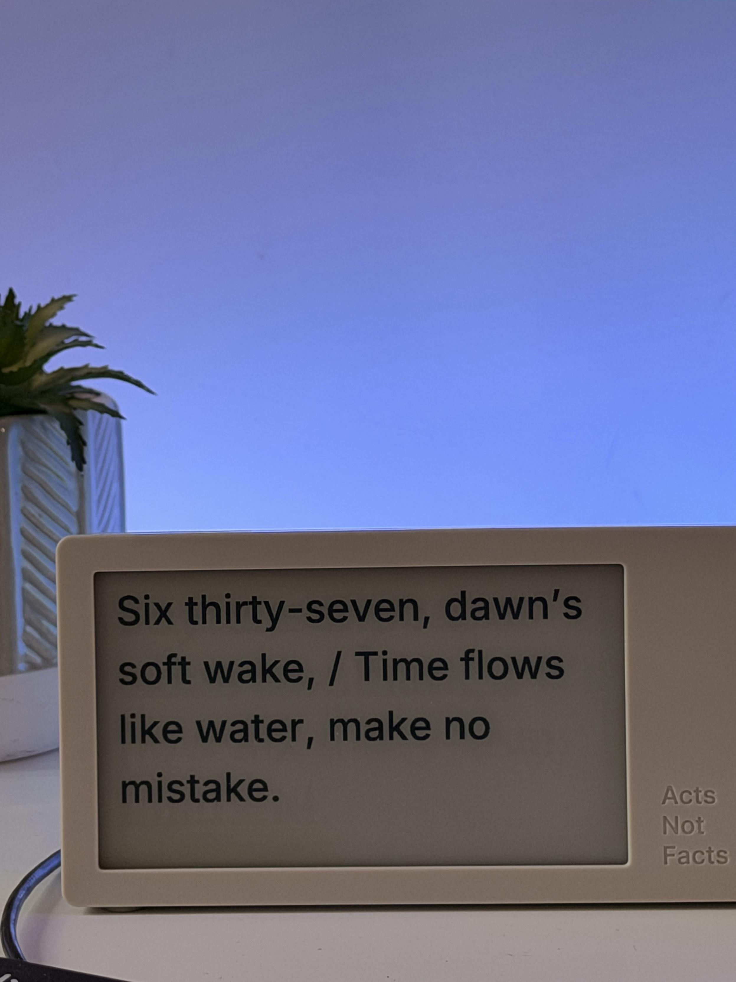 A digital display shows the text: "Six thirty-seven, dawn's soft wake, / Time flows like water, make no mistake." The display is positioned on a desk beside a small potted plant, with a softly lit background.