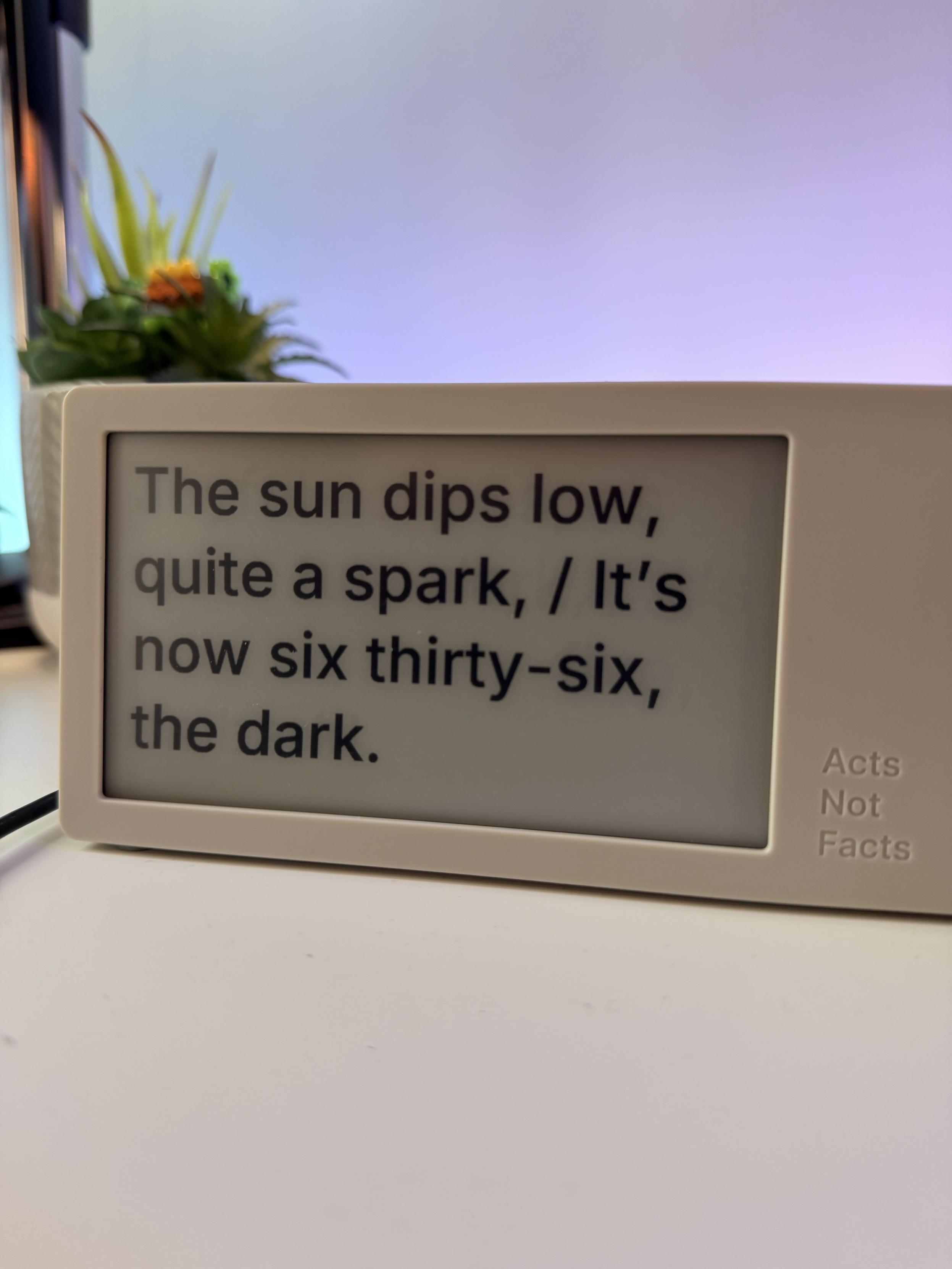 The image features a display screen showing a poetic text: "The sun dips low, quite a spark, / It's now six thirty-six, the dark." A small potted plant is visible in the background.