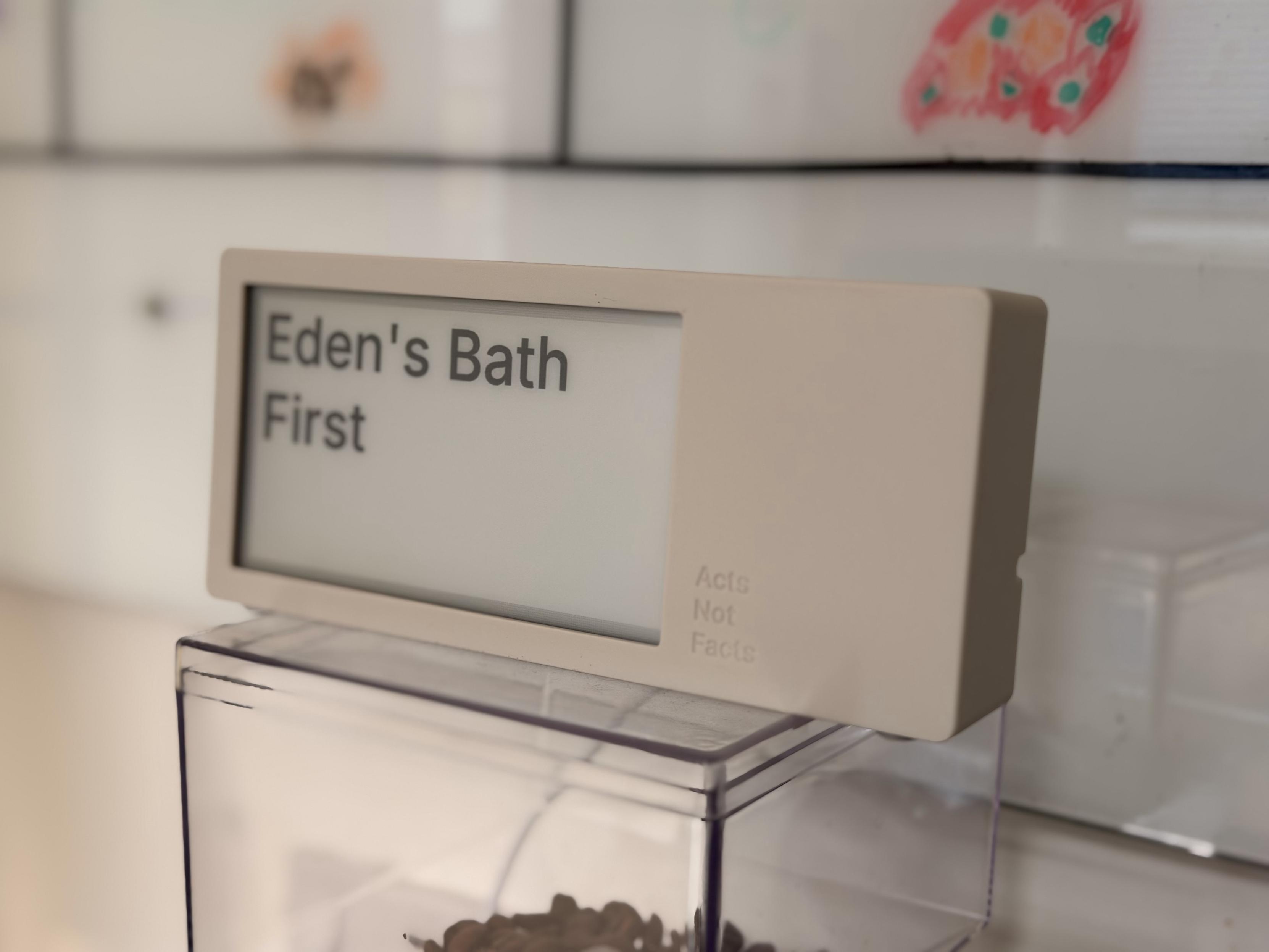 A white display sign with the text "Eden's Bath" and "First" is visible. The sign is positioned above a clear container filled with small items. The background features blurred colorful artwork.