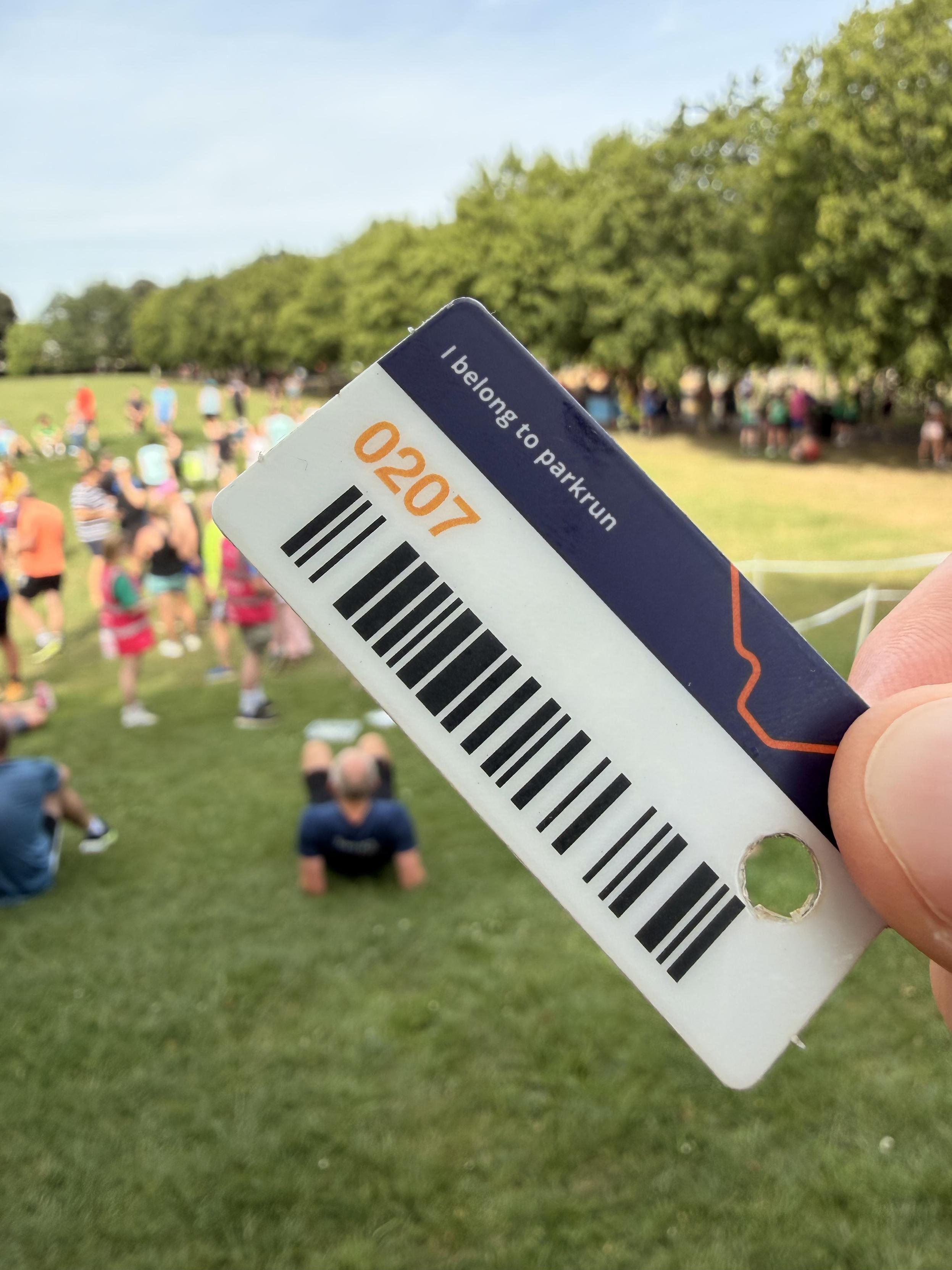 A hand holds a park run barcode card displaying the text "I belong to parkrun" and a barcode. In the background, a crowd participates in an outdoor event, surrounded by greenery and visible runners.